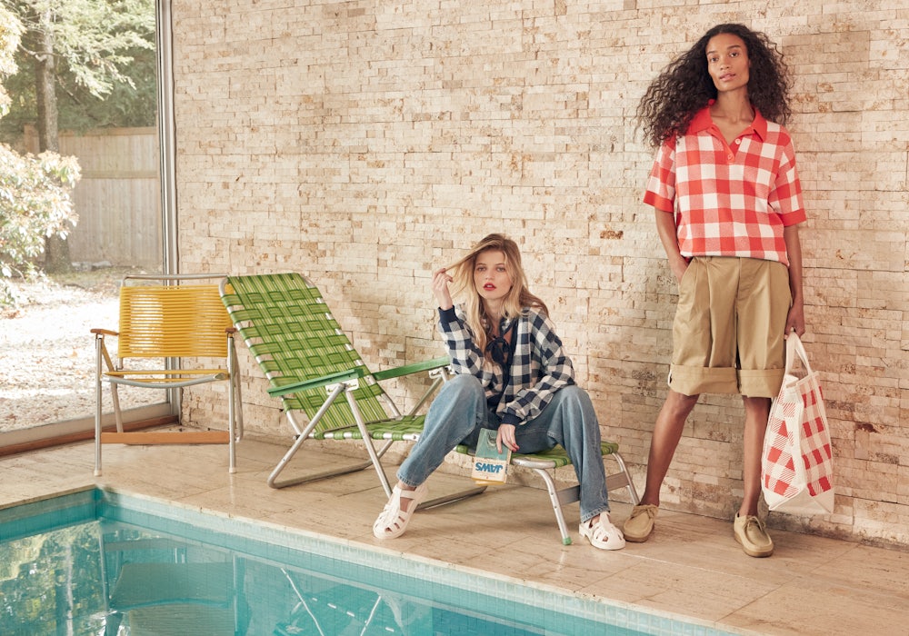 KULE- Two female models sitting by pool. One wearing navy Louise gingham cardigan and one wearing the Avril Gingham poppy sweater.