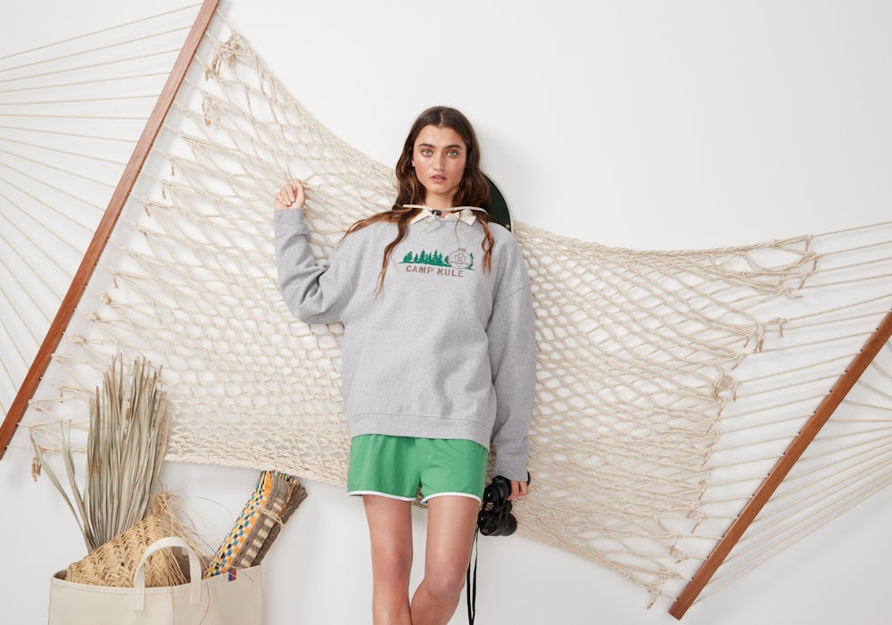 KULE- KULE model wearing Camp KULE Raleigh Sweatshirt, paired with The Short in green. She is leaning against a hammock.