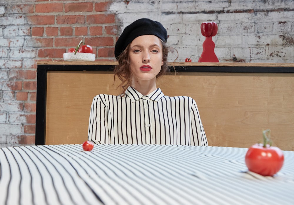 KULE- Model wearing black and white stripe silk blouse, wearing a black french hat, sitting at a table with a matching black/white stripe tablecloth. It's all very French.