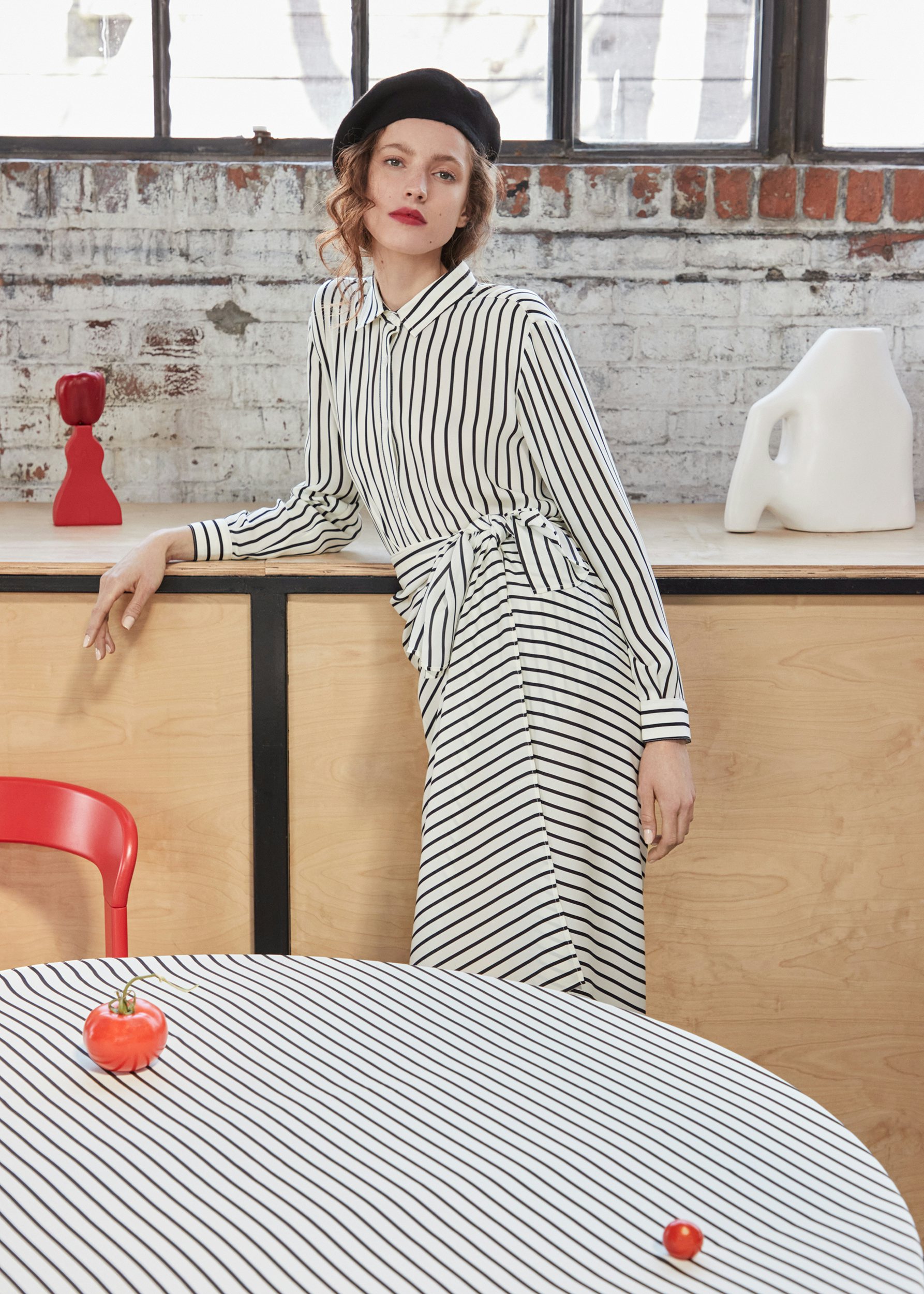 Model wearing the KULE Ponza black and white stripe silk top, with matching Elba black and white stripe wrap skirt. She is wearing a black beret and leaning on a counter. It's all so very French-inspired.