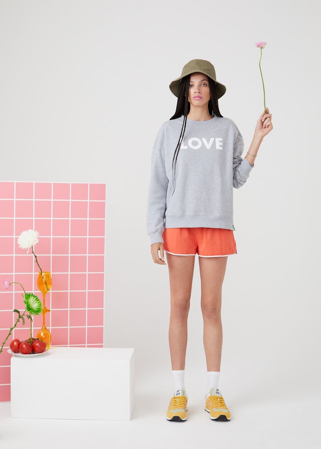 KULE Green - Model wearing LOVE organic cotton sweatshirt holding a pink flower next to a block with a flower in a vase and a plate of fruit