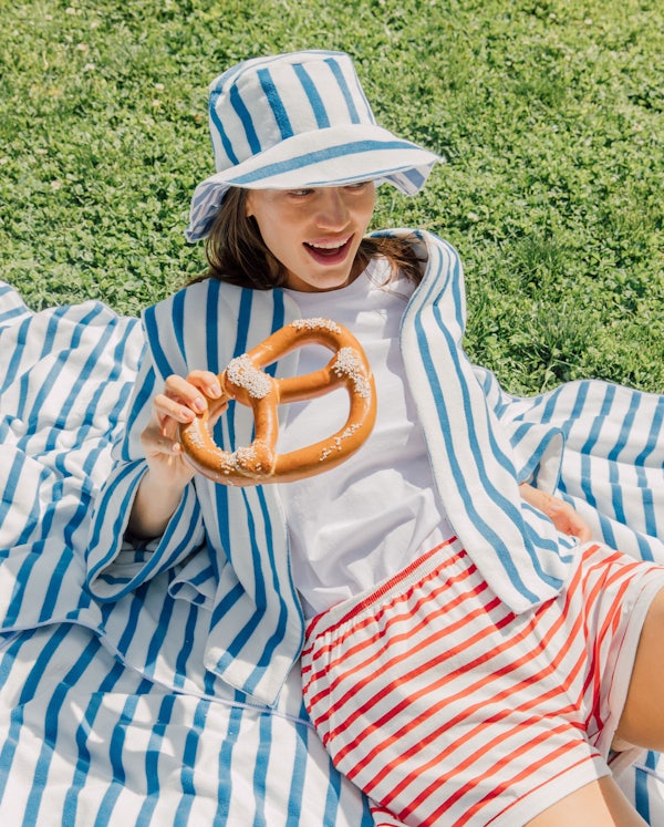 Model sititng on a beach towel in the grass wearing a hat and jacket from the LilyEve collection while eating a soft pretzle.